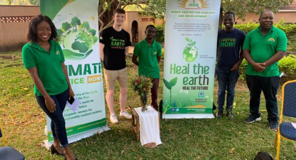 Sam, Martha and Brother ngoni stand outside beside Laudato si banners under the shade of a tree.