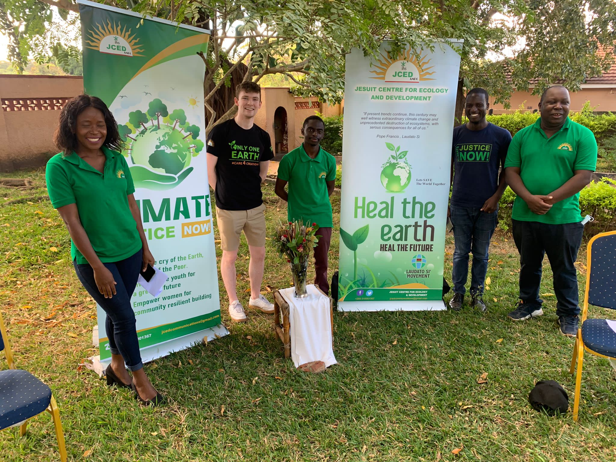 Sam, Martha and Brother ngoni stand outside beside Laudato si banners under the shade of a tree.