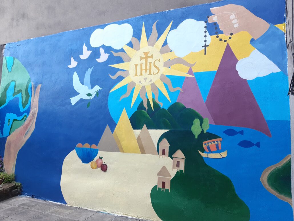 completed mural. it depicts hills and mountains. hands cradling planet earth. Hill and mountains, a blue sky and the ocean. the dove of peace is above the landscape scene.