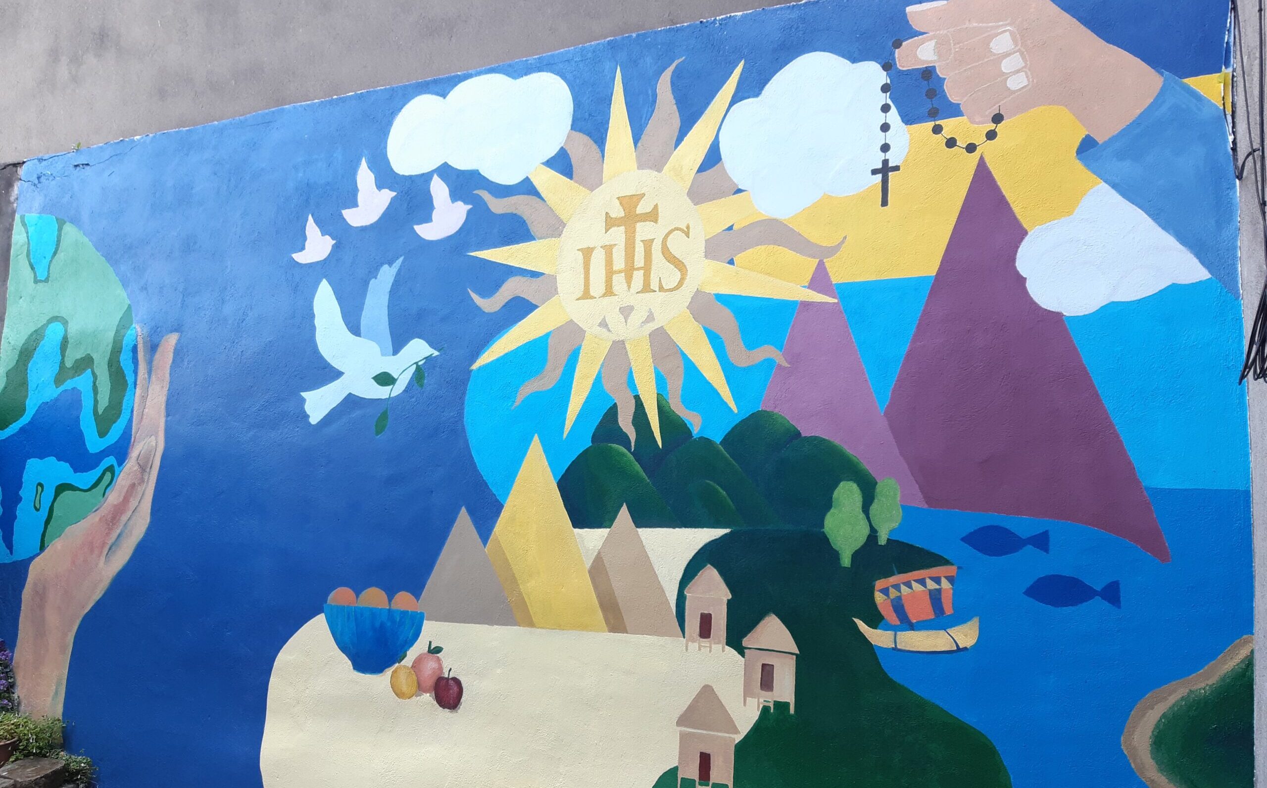 completed mural. it depicts hills and mountains. hands cradling planet earth. Hill and mountains, a blue sky and the ocean. the dove of peace is above the landscape scene.
