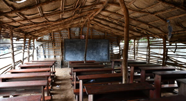 classroom hun made from sticks and sheeted coverings with wooden desks, sand floor. A blackboard stands at the front of the room.