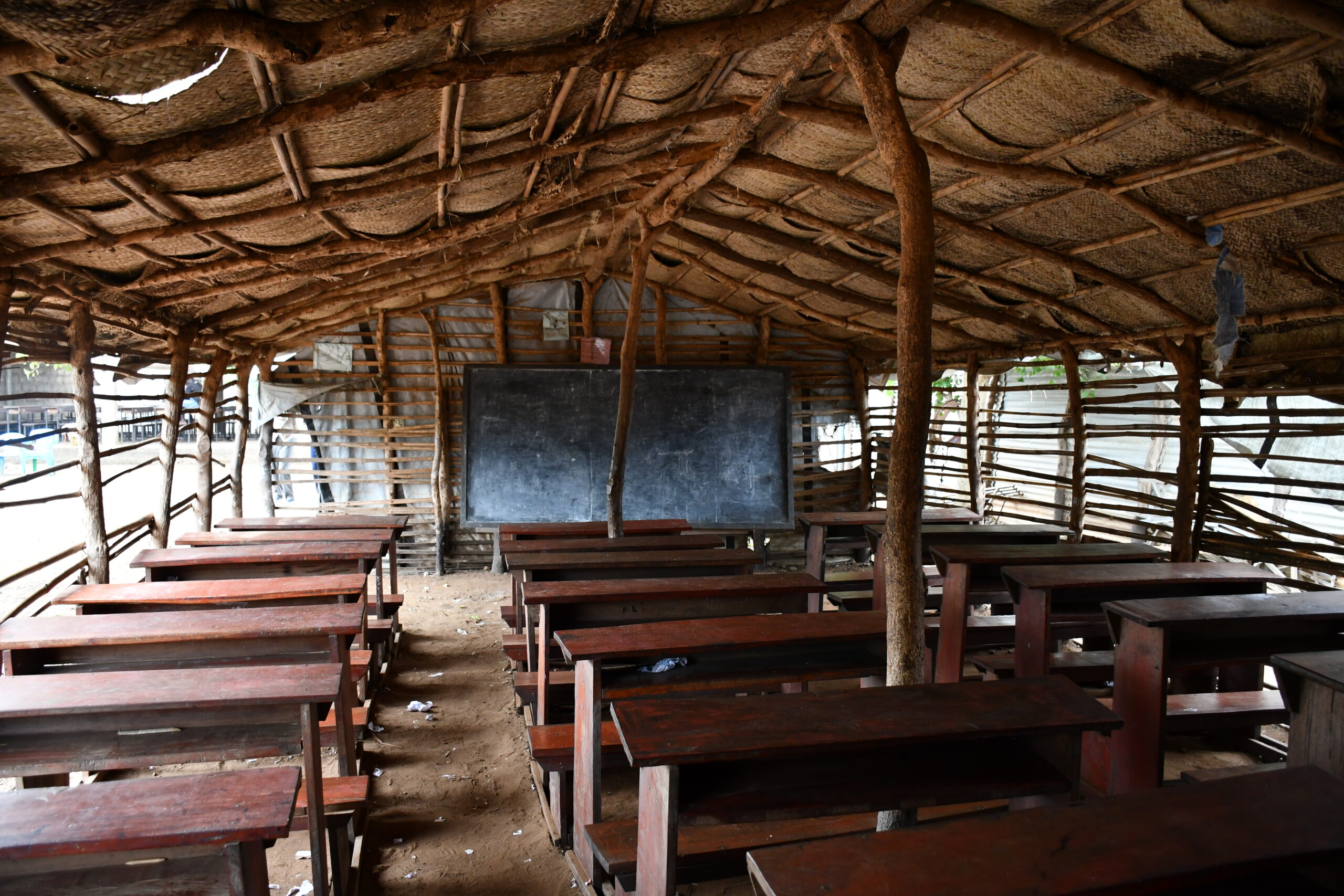 classroom hun made from sticks and sheeted coverings with wooden desks, sand floor. A blackboard stands at the front of the room.