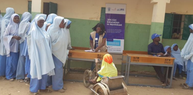 Shatu, a young girl in a wheelchair is registering for school. other school girls are in line to register behind Shatu. 