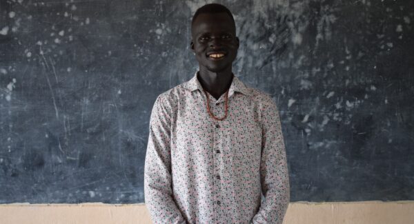 Gabriel, a teacher at SPF smiles at the camera. behind him is a blackboard