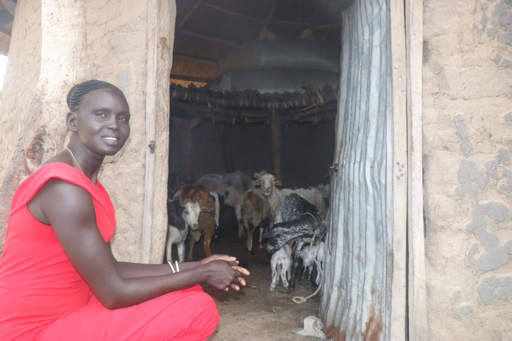 Atuenyd, a woman farmer smiles at the camera. She is showing the viewer her goats inside a tukul.