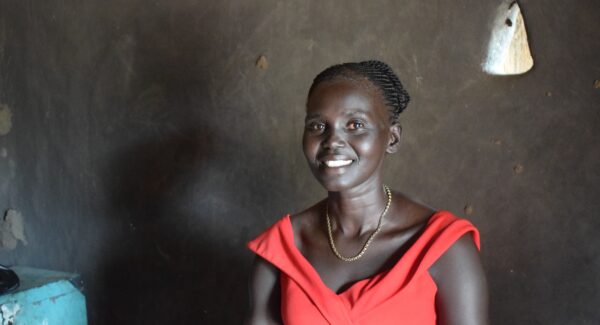 A woman farmer, Atuenydelg smiles at camera. She is sat in her tukul posing for photo.