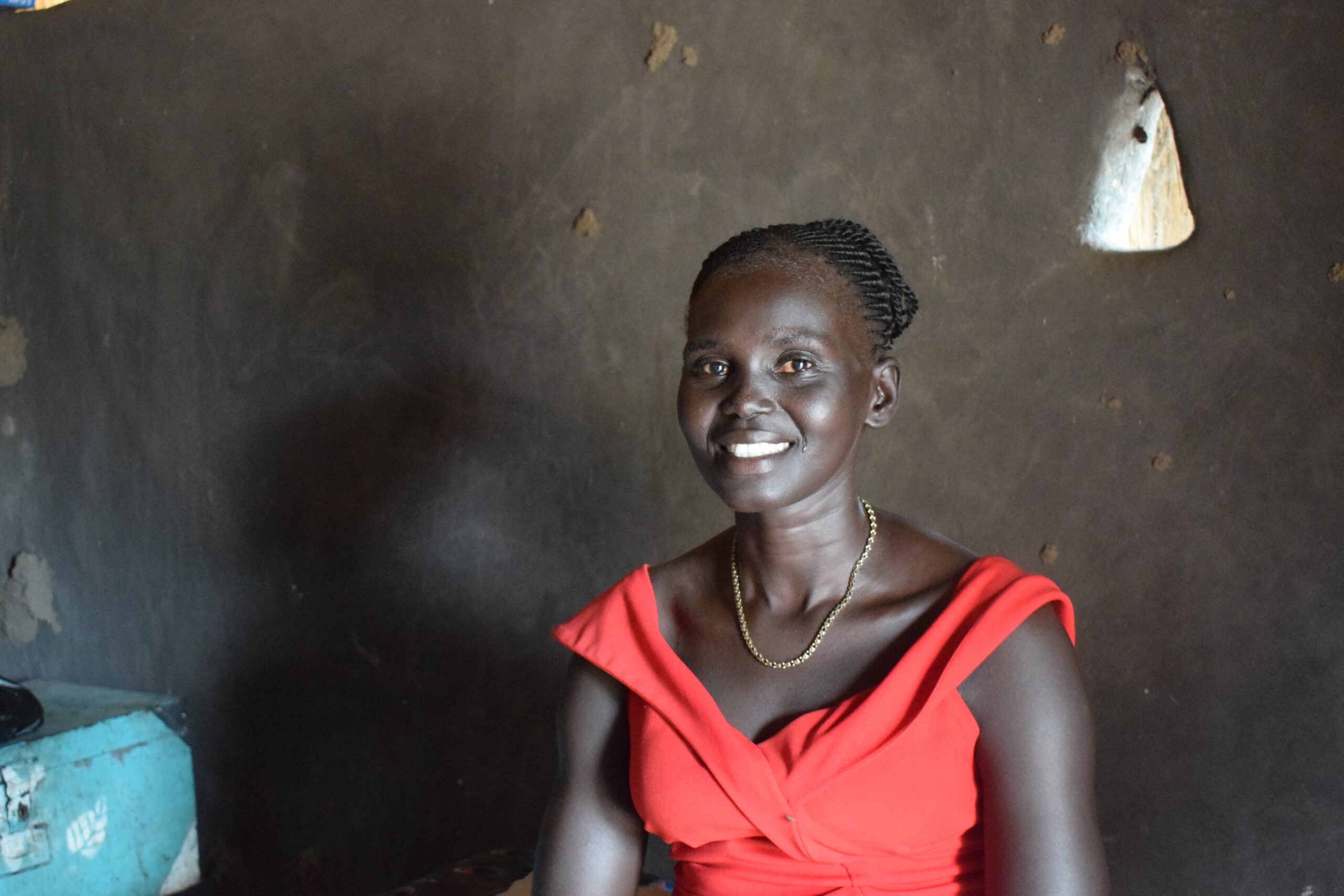 A woman farmer, Atuenydelg smiles at camera. She is sat in her tukul posing for photo.