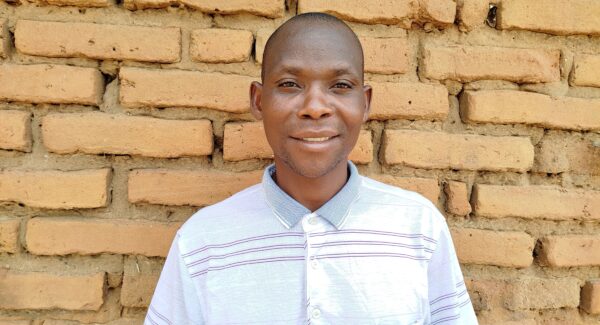 Happy, A Malawian farmer smiles at the camera. he stands with a brick wall behind him and wears a white shirt.
