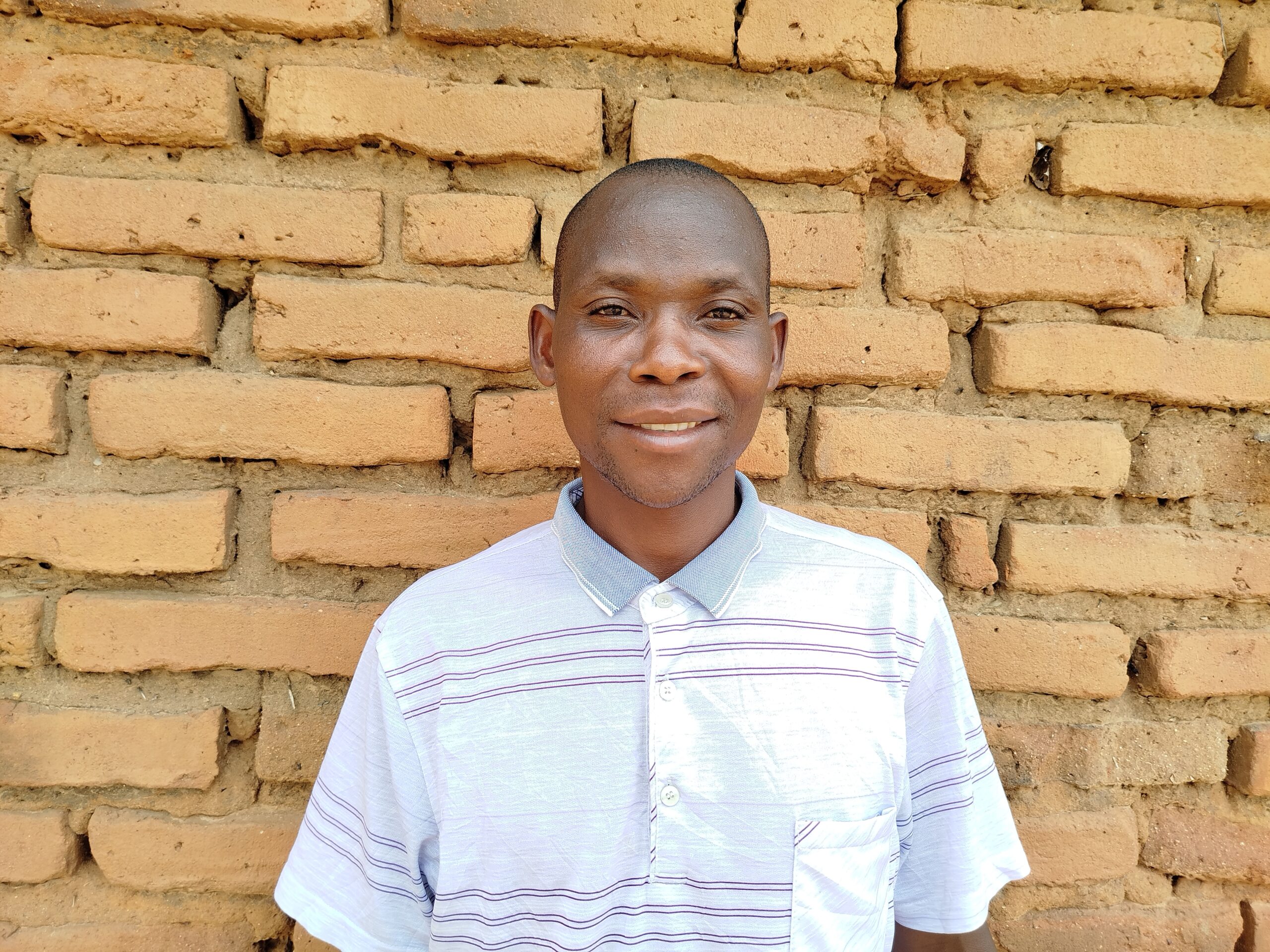 Happy, A Malawian farmer smiles at the camera. he stands with a brick wall behind him and wears a white shirt.