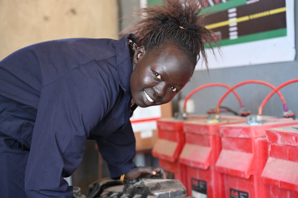 Martha wears navy overalls and stands over a car engine