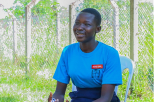 A school girl is smiling sat outside the school yard. she is wearing a school uniform and a badge that reads: 'head girl'.