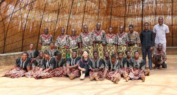Timmayo is posing with a cultural troupe in a large grass hut. They all smile at the camera.