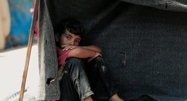 a boy sits under makeshift tent in Gaza. he looks toward the camera pensively.