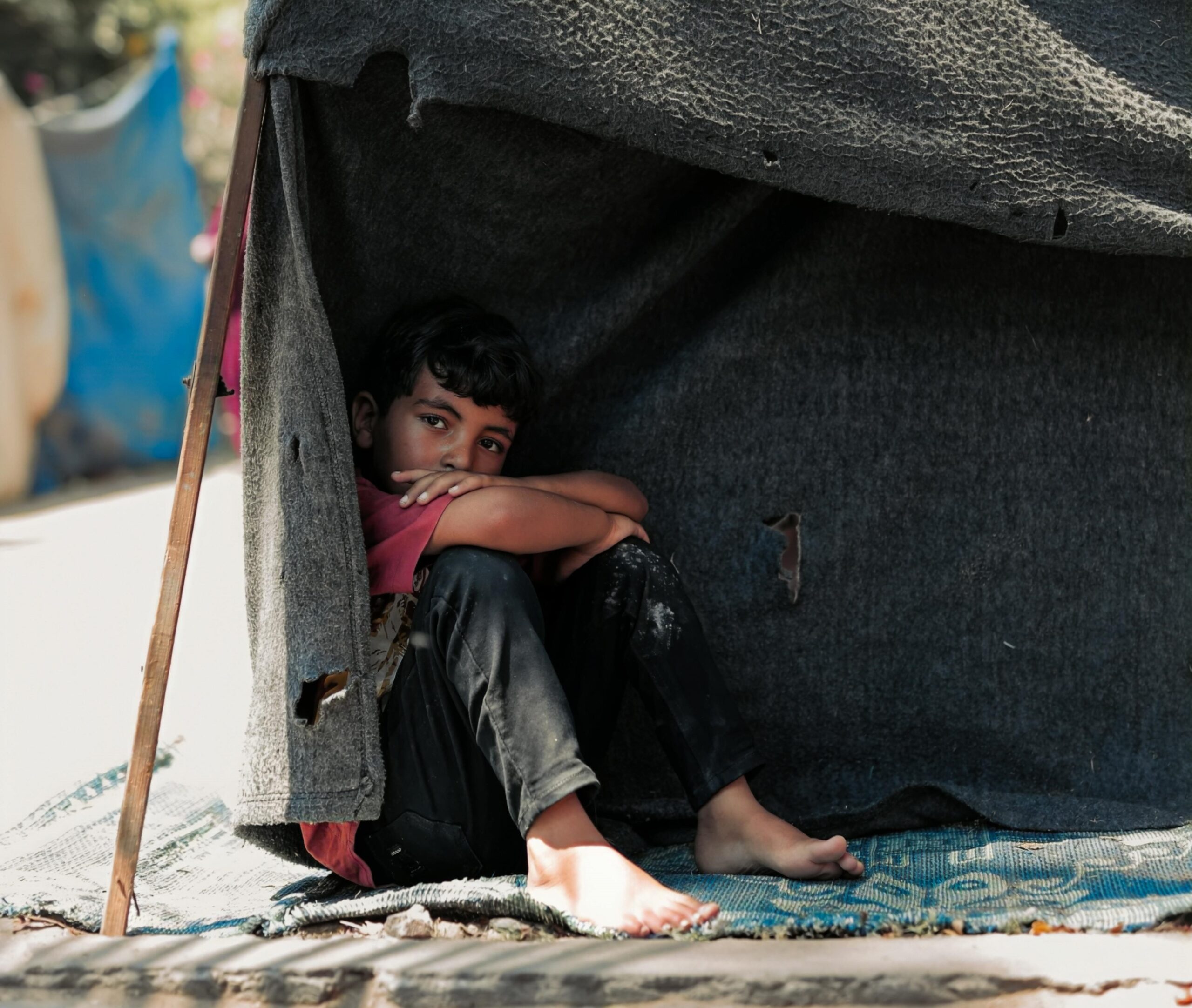 a boy sits under makeshift tent in Gaza. he looks toward the camera pensively.