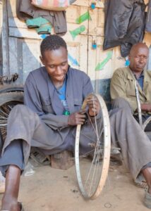 Joseph sits on the floor mending the spokes of a bicycle wheel. He is smiling shyly. 