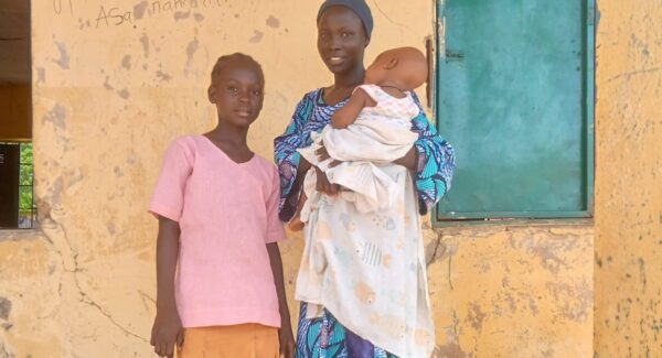 Evelyn poses outside the classroom block with her mother and baby sibling. they smile toward the camera.