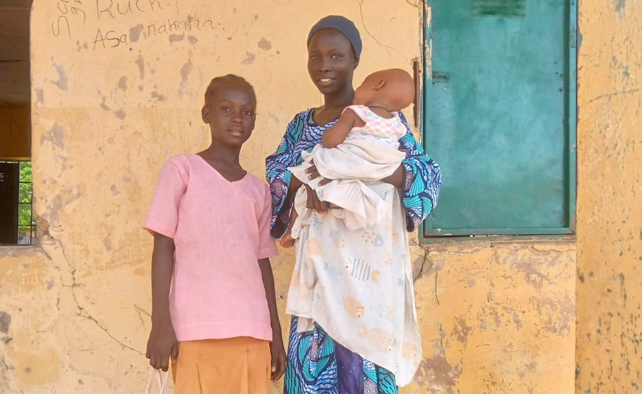Evelyn poses outside the classroom block with her mother and baby sibling. they smile toward the camera.