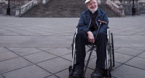Sean sits in a wheelchair smiling toward the camera. He is wearing a cap and glasses.