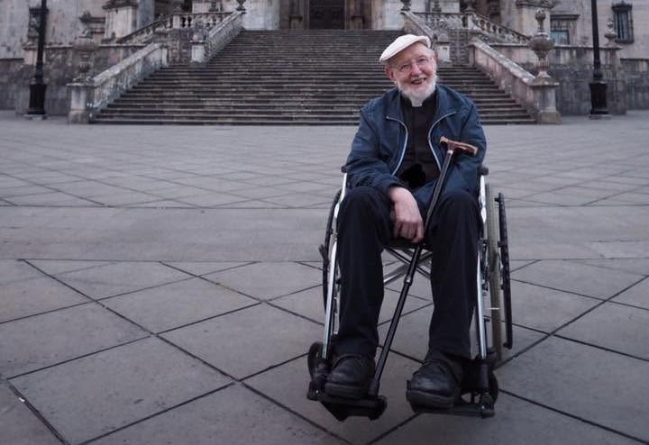 Sean sits in a wheelchair smiling toward the camera. He is wearing a cap and glasses.