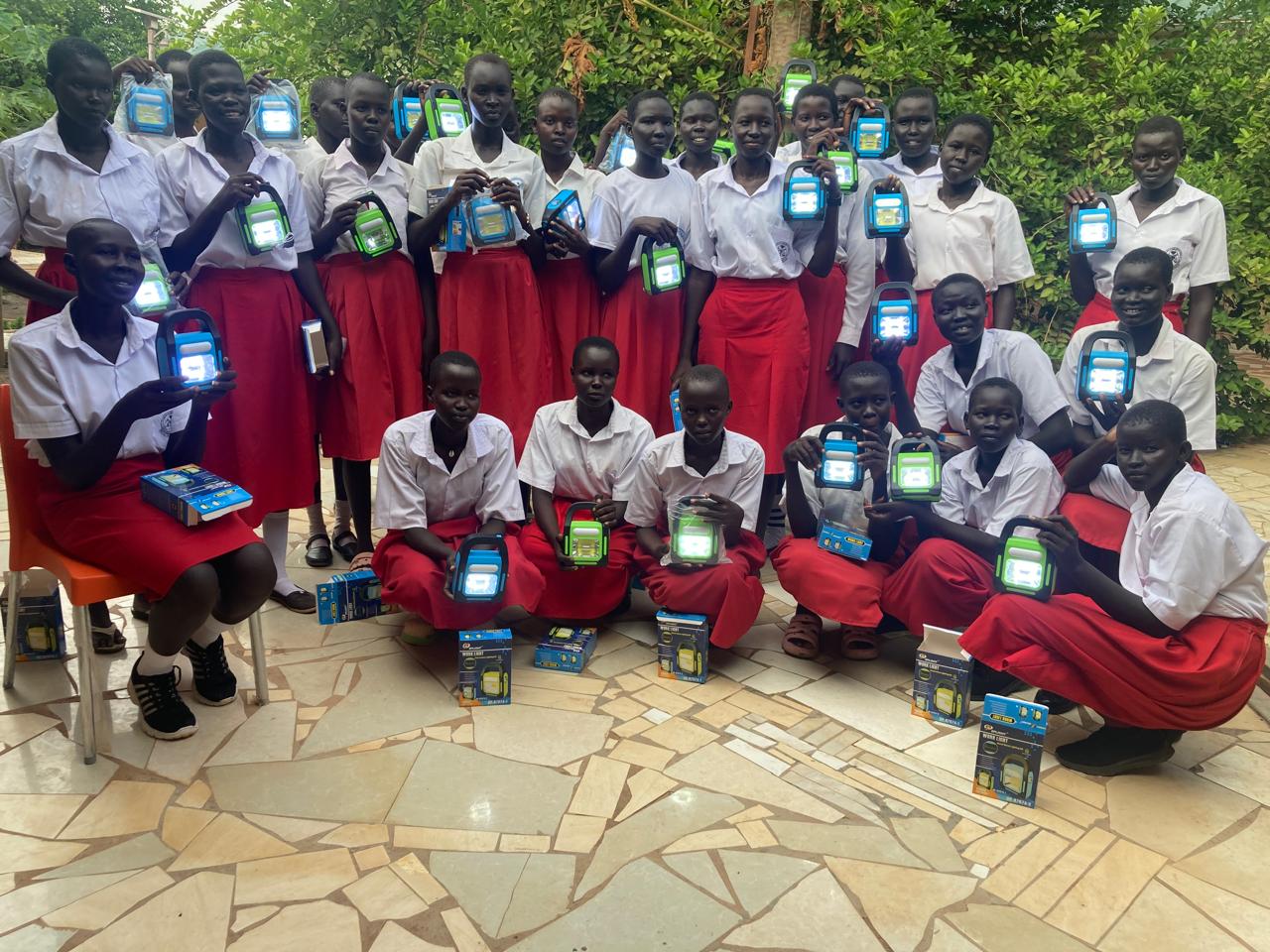 a group of students in uniform smile at the camera. they are holding their solar lamps