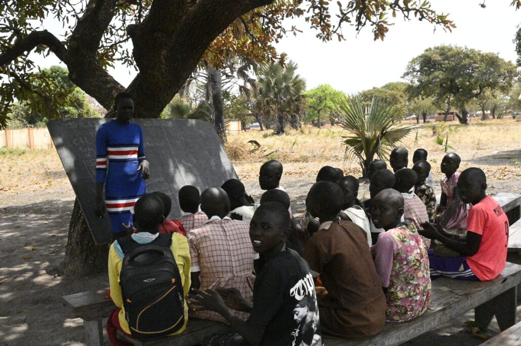 Monica teaches at a blackboard which is resting against a tree trunk. she s teaching a class under the shade of a tree.