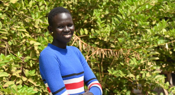 Monica, teacher at St Justina primary in South Sudan poses outside. she is smiling toward the camera and wears a blue dress.