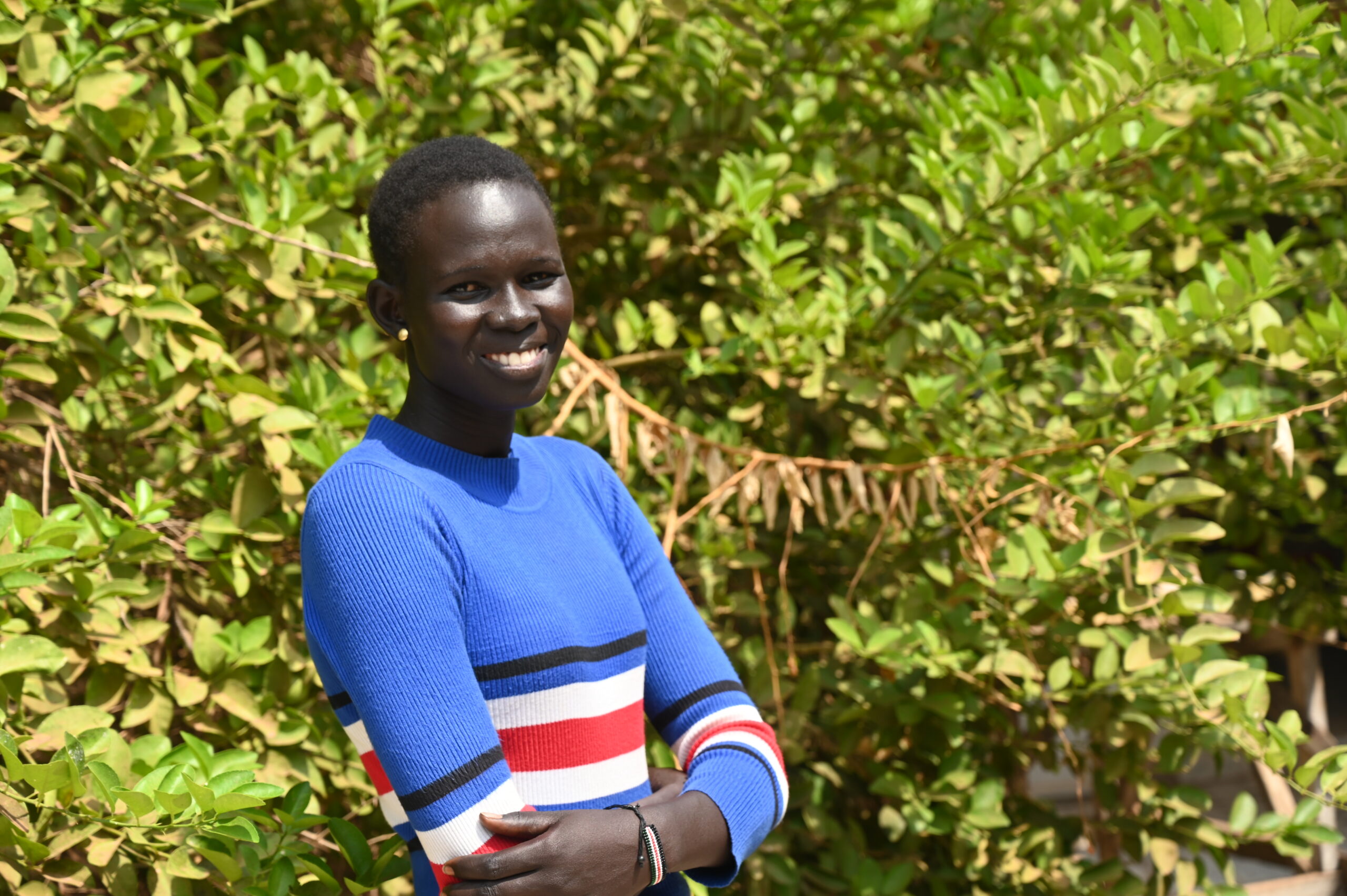 Monica, teacher at St Justina primary in South Sudan poses outside. she is smiling toward the camera and wears a blue dress.